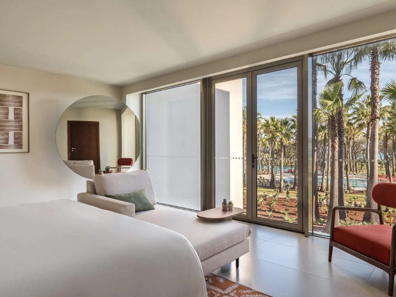 A modern hotel room with a large bed, chaise lounge, and a red armchair, featuring a floor-to-ceiling window with a view of palm trees and a swimming pool.