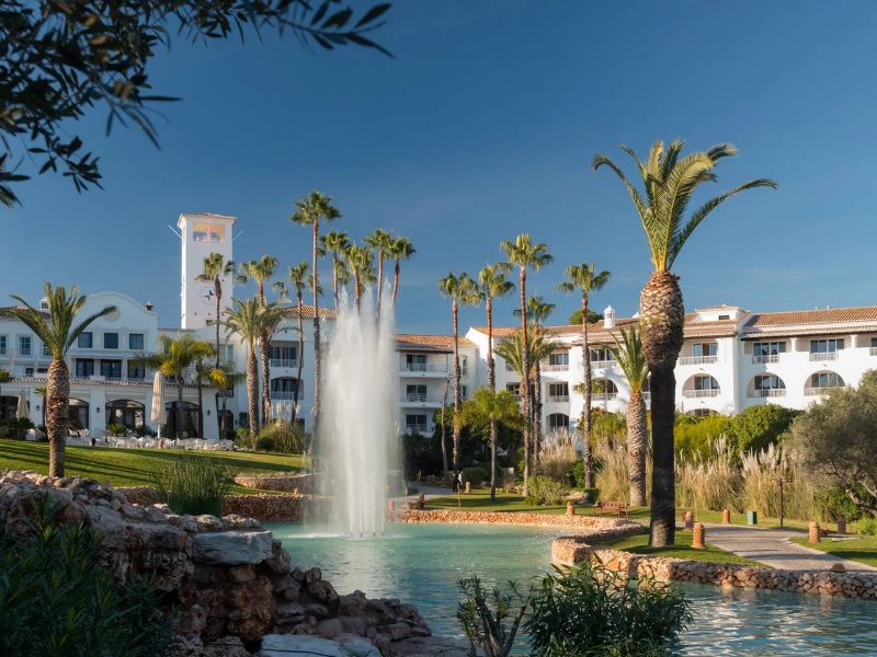 A white hotel with a tower, surrounded by palm trees and a water fountain in a pond, under a clear blue sky.