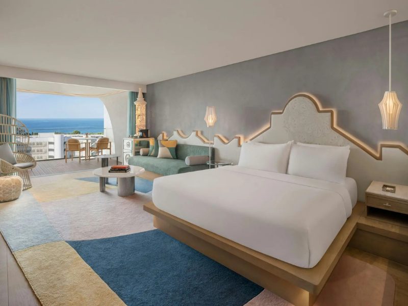 A spacious hotel suite with a large bed, a green sofa, and a balcony overlooking the ocean. The room features a colorful geometric rug and modern lighting fixtures.