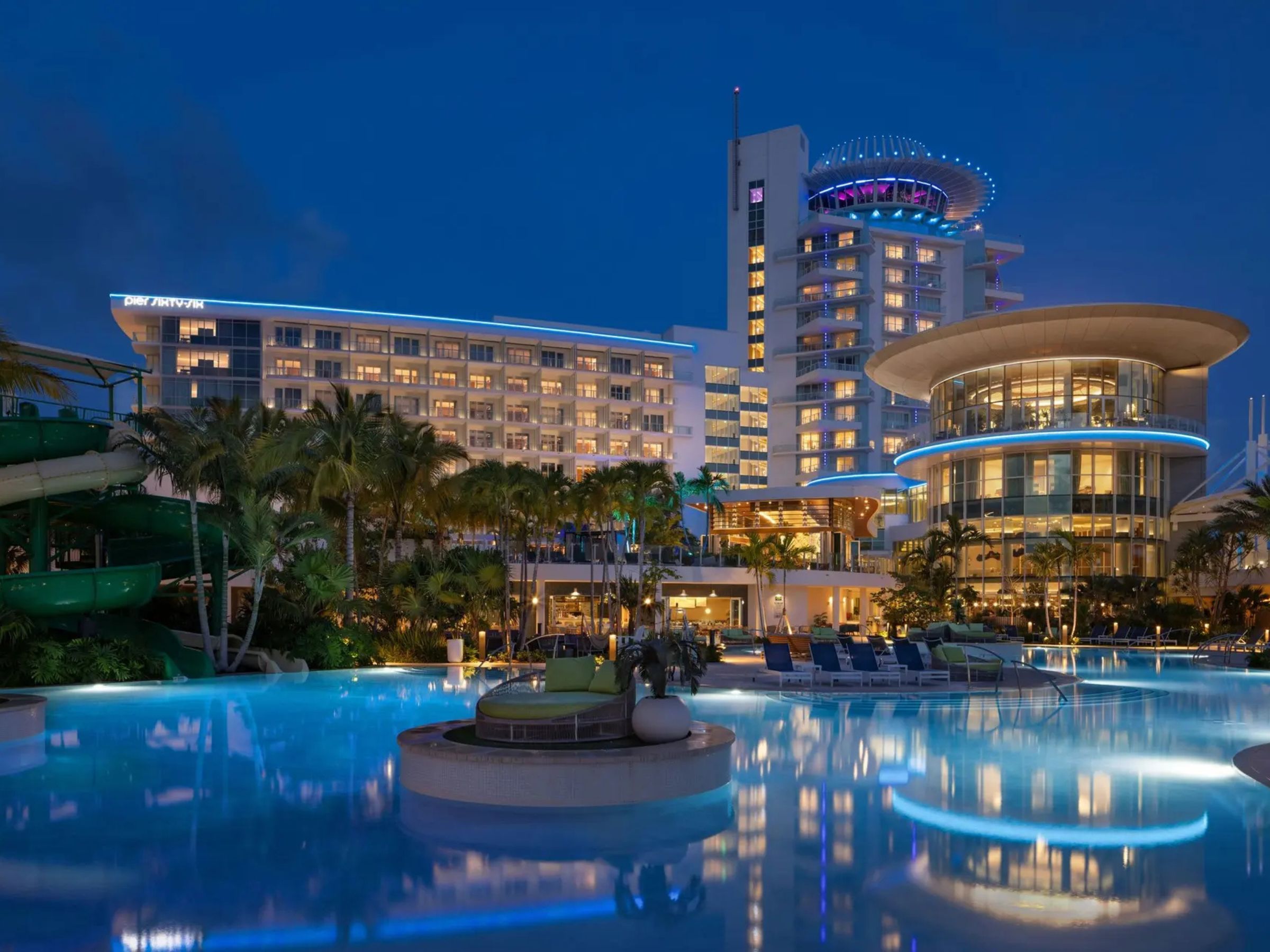Luxury resort illuminated at twilight, featuring a sprawling lagoon-style swimming pool, palm trees, waterslides, and contemporary hotel towers with vibrant architectural lighting.